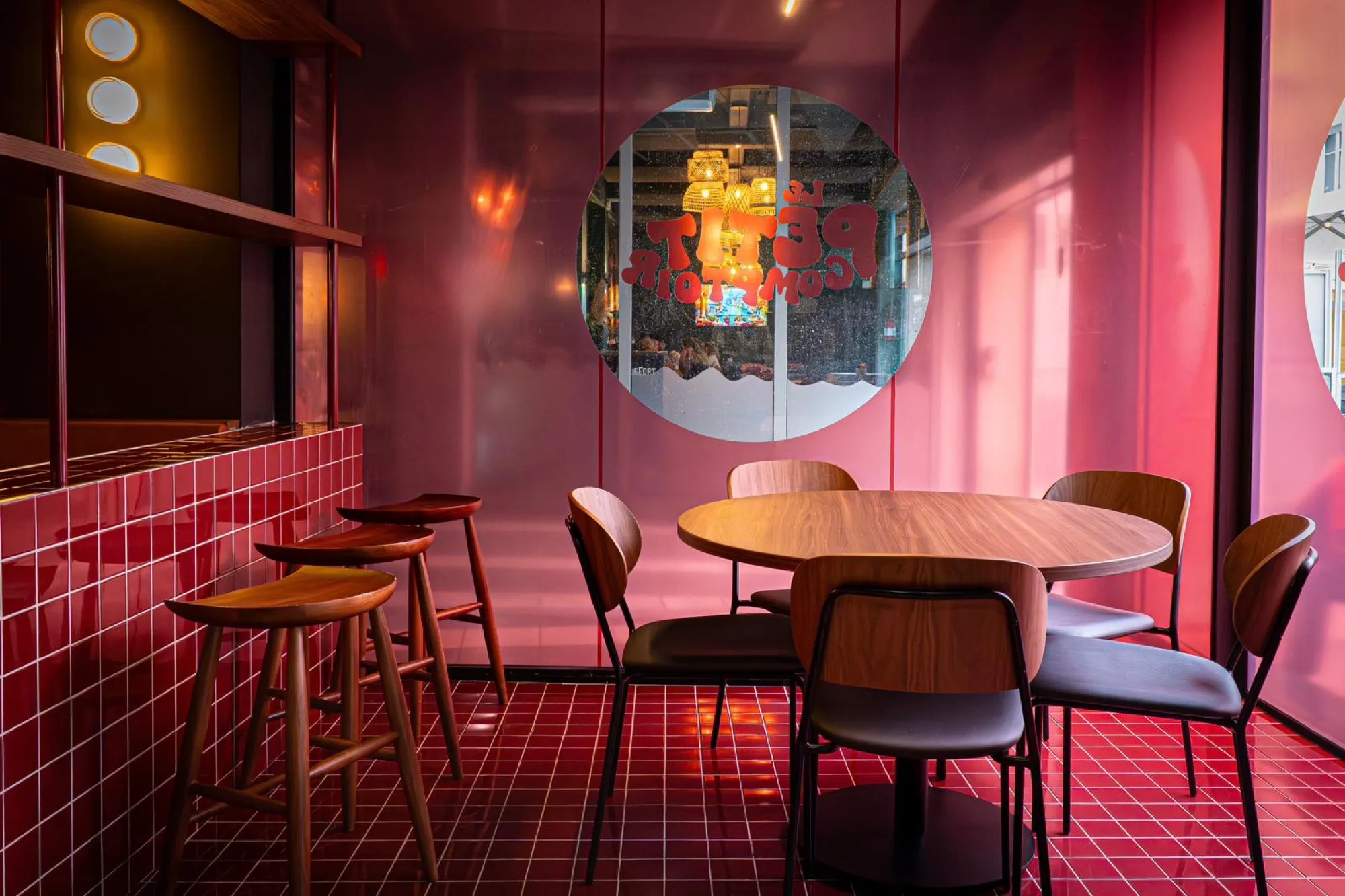 Salle du bar Le Petit Comptoir à Lille : murs laqués rouge et rose, sol et bar en carrelage rouge, table ronde et tabourets en bois, hublot vitré portant le nom du bar.